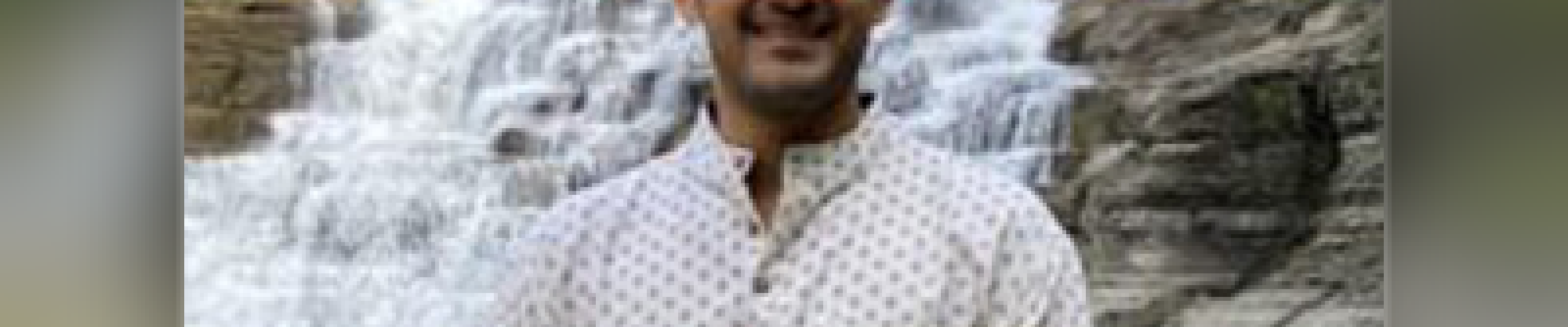 A color photo of a man standing outside with a waterfall in the background