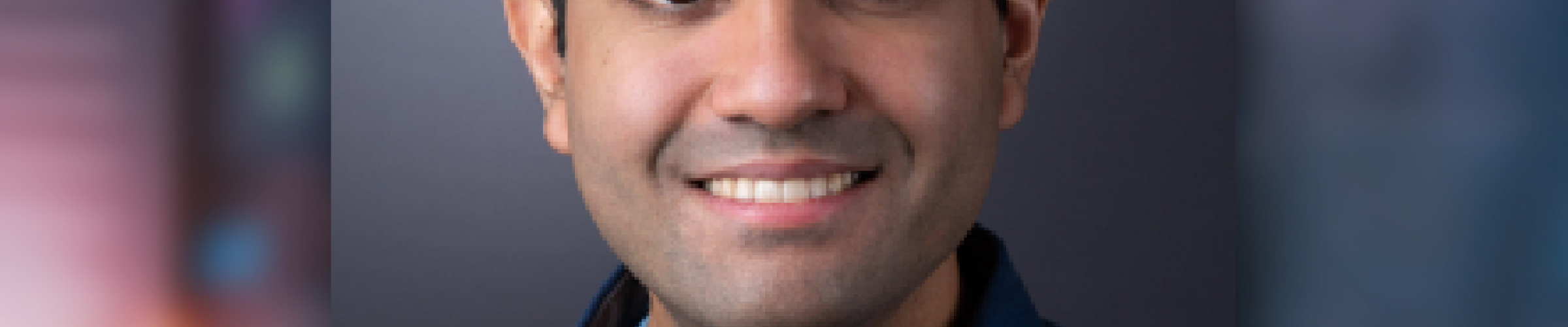 A color photo of a man smiling for a photo with a blurred blue background