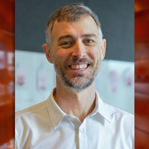 A color photo of a man smiling for a photo with a blurred red background