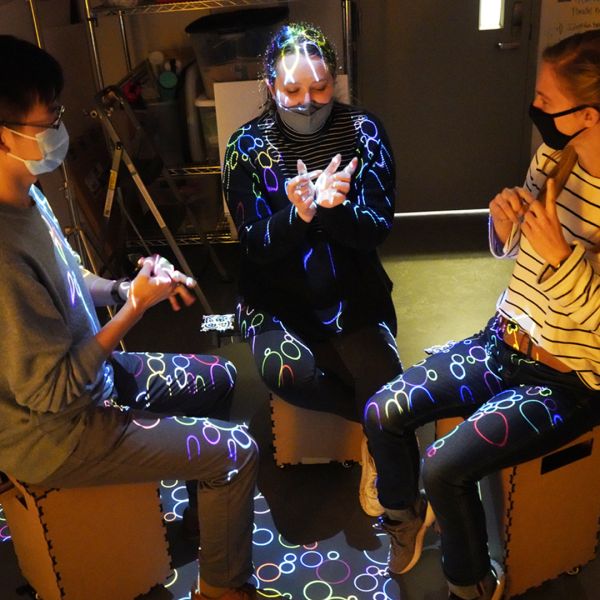 A color photo of 3 young people participating in a science experiment with colorful, circular lights