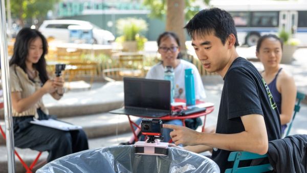 A color photo showing doctoral student Frank Bu, foreground, checks the camera on one of the trashbots during deployment in Albee Square, Brooklyn, in July. 