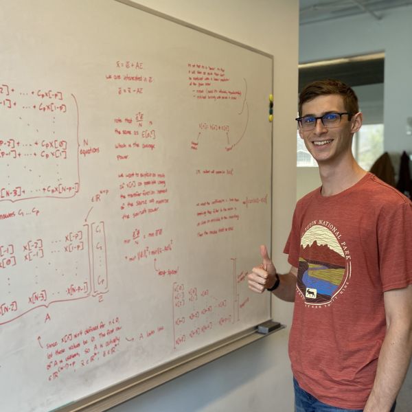 A color photo of a man standing beside a chalkboard with his thumbs up