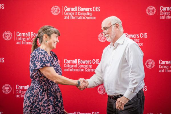 A color photo of a man and woman shaking hands in front of a Cornell Bowers CIS, red backdrop