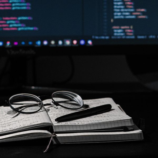 Notebooks in front of a computer monitor, which shows computer code. // Photo by Kevin Canlas courtesy of Unsplash