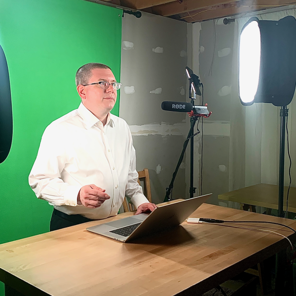 Color photo of a man in a white shirt and glasses being recorded in a studio