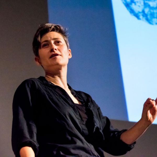 A color photo showing a woman talking in front of a large video screen