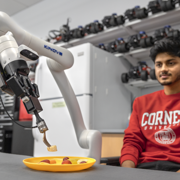 A color photo of a man in a red Cornell sweatshirt sitting in front of a robotic arm